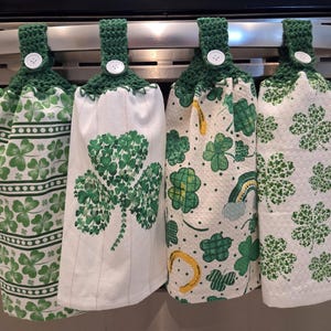 May include: Four green and white kitchen towels with crochet tops. Each towel features a different St. Patrick's Day design, including shamrocks and a rainbow. The towels are hanging on a metal bar.
