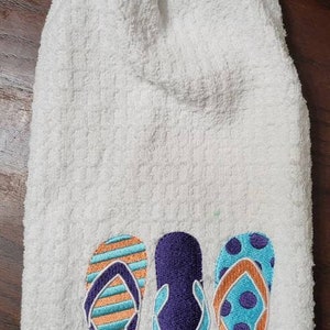 May include: White kitchen towel with a blue crocheted top and a button. The towel has three embroidered flip flops in orange, purple, and blue.