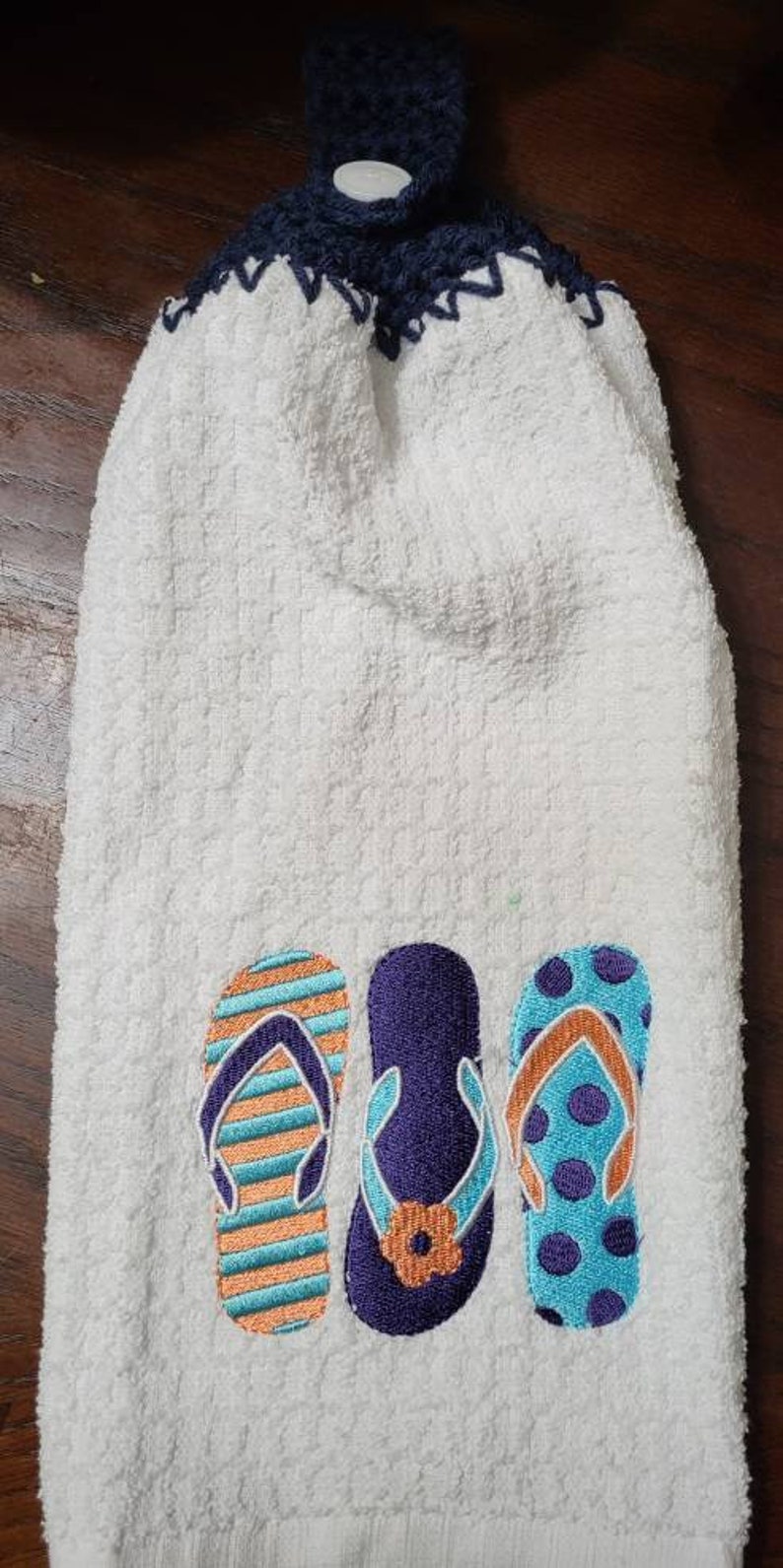 May include: White kitchen towel with a blue crocheted top and a button. The towel has three embroidered flip flops in orange, purple, and blue.