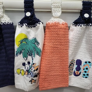 May include: Four kitchen towels with crocheted loops and buttons. The towels feature different designs, including a palm tree with Mickey Mouse and friends, flip flops, and a solid orange color.