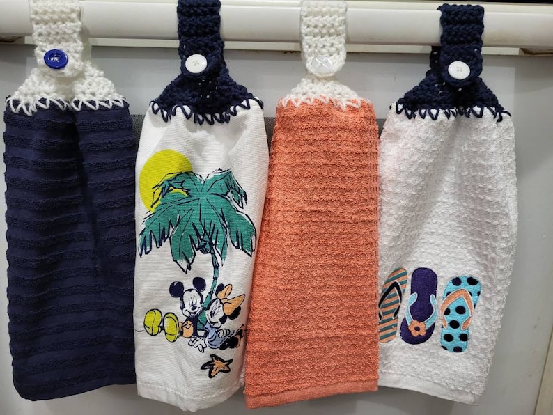 May include: Four kitchen towels with crocheted loops and buttons. The towels feature different designs, including a palm tree with Mickey Mouse and friends, flip flops, and a solid orange color.