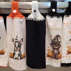 May include: Five Halloween-themed kitchen towels with crocheted loops and buttons. Two towels feature a black and white illustration of a French bulldog wearing a witch's hat and holding a pumpkin. Two towels feature a black cat wearing a witch's hat and holding a pumpkin. One towel is black with a white crocheted border.