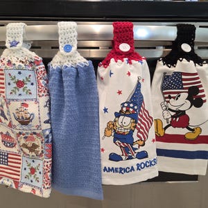 May include: Four hanging kitchen towels with crochet tops. One towel features a patchwork design with patriotic motifs. Another is solid blue. The third has Garfield with an American flag, and the last has Mickey Mouse with a flag.