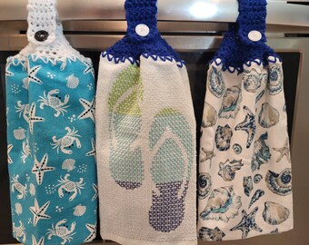May include: Three kitchen towels with crocheted loops and buttons. The towels have a nautical theme with images of seashells, starfish, and crabs. The towels are hanging on a silver oven rack.