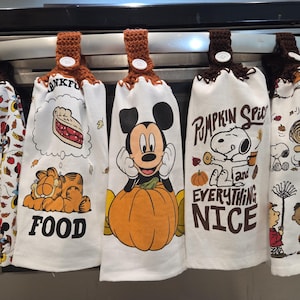 May include: Five white kitchen towels with brown crochet tops hang from a metal bar. The towels feature cartoon characters like Minnie Mouse, Garfield, and Mickey Mouse with a pumpkin. Other towels have text that says "FOOD" and "Pumpkin Spice and Everything Nice."