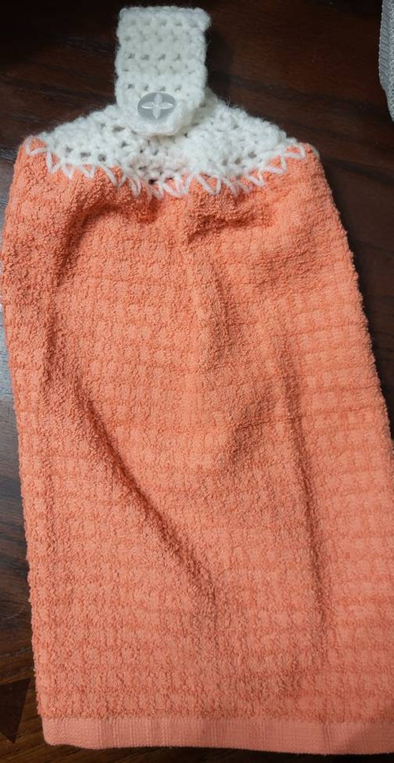 May include: A peach-colored kitchen towel with a white crocheted loop and button at the top.