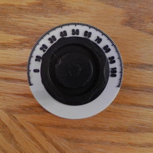May include: A white and black dial with numbers from 0 to 100. The dial has a black center and a white outer ring with black markings. The dial is on a wooden surface.