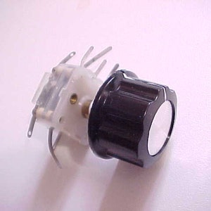 May include: A black and white knob with a clear plastic base and metal contacts. The knob has a white center and a black outer ring with ridges.