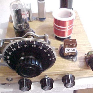 May include: A close-up of a vintage radio kit with a wooden base, a dial with numbers from 0 to 100, a black knob, and a red coil. The kit also includes a vacuum tube, a capacitor, and a resistor.