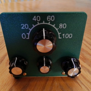 May include: A dark green control panel featuring a large central knob positioned above a curved scale marked from 0 to 100. Three smaller black knobs with silver centers are arranged in a row along the bottom edge of the rectangular device.