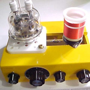 May include: A yellow box with four knobs and a clear glass tube with a white base. The tube has a red and white coil on top of it. The box is a radio transmitter.