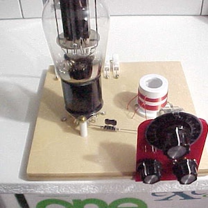 May include: A close-up of a vintage vacuum tube radio circuit with a large vacuum tube, a coil with red wire windings, and three knobs on a red panel.