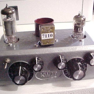 May include: A vintage silver radio with three knobs and two glass vacuum tubes. The radio has a black label that reads "KWT" and a smaller label with the number "7110".