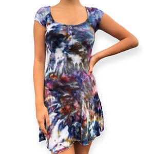 Pode incluir: Vestido de gola redonda e manga curta com estampa tie-dye em tons de azul, roxo, vermelho e branco. O vestido tem uma frente abotoada e uma saia rodada. O tecido parece ser malha canelada.