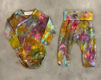 Puede incluir: Un conjunto de body cruzado de manga larga y pantalón a juego con un vibrante estampado tie-dye en tonos amarillo mostaza, magenta y azul verdoso. El body tiene una etiqueta blanca con el texto "TOCAYO" y cierres a presión.