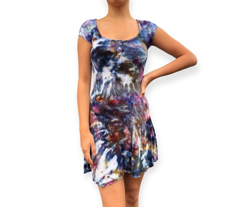 Pode incluir: Vestido de gola redonda e manga curta com estampa tie-dye em tons de azul, roxo, vermelho e branco. O vestido tem uma frente abotoada e uma saia rodada. O tecido parece ser malha canelada.