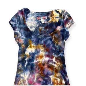 Pode incluir: Uma camiseta de manga curta com decote redondo e um padr&atilde;o tie-dye em tons de azul, roxo, amarelo e rosa. A camisa tem uma carcela com bot&otilde;es e uma pequena etiqueta perto da gola.