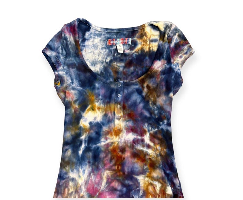 Pode incluir: Uma camiseta de manga curta com decote redondo e um padr&atilde;o tie-dye em tons de azul, roxo, amarelo e rosa. A camisa tem uma carcela com bot&otilde;es e uma pequena etiqueta perto da gola.