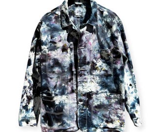 May include: A tie-dye denim jacket with a button-up front, four front pockets, and white stitching. The jacket features a unique pattern of blues, purples, and white, creating a marbled effect. The jacket is hanging on a black hanger.