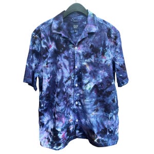May include: A short-sleeved button-down shirt with a tie-dye pattern in shades of blue, purple, and pink. The shirt has a chest pocket and the GAP logo on the collar. The shirt is hanging on a black hanger.