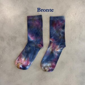 Puede incluir: Un par de calcetines de media ca&ntilde;a con un patr&oacute;n de te&ntilde;ido anudado en tonos azul profundo, morado y toques de rosa. La palabra Bronte est&aacute; impresa en azul oscuro sobre los calcetines. Tienen un pu&ntilde;o acanalado y una textura de punto lisa.