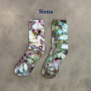 Puede incluir: Un par de calcetines de media ca&ntilde;a con un patr&oacute;n de te&ntilde;ido anudado en tonos blanco, rosa, verde y marr&oacute;n. La palabra "Siena" est&aacute; impresa en azul sobre los calcetines. Los calcetines tienen un pu&ntilde;o acanalado y un aspecto suave y mezclado.