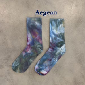 Puede incluir: Un par de calcetines de media ca&ntilde;a con un patr&oacute;n tie-dye en tonos azul, verde y magenta. La palabra Aegean est&aacute; impresa en azul oscuro sobre los calcetines. Los calcetines tienen un pu&ntilde;o acanalado y un aspecto suave y difuminado.