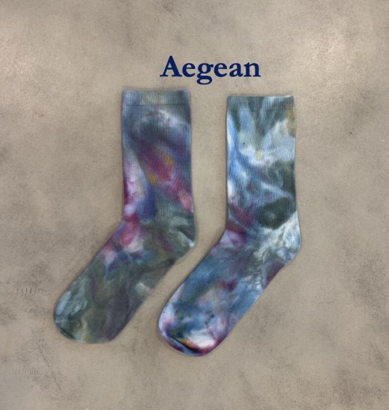 Puede incluir: Un par de calcetines de media ca&ntilde;a con un patr&oacute;n tie-dye en tonos azul, verde y magenta. La palabra Aegean est&aacute; impresa en azul oscuro sobre los calcetines. Los calcetines tienen un pu&ntilde;o acanalado y un aspecto suave y difuminado.