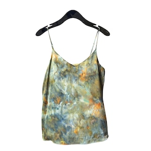Puede incluir: Un camisola teñida con un diseño tie-dye con cuello en V y tirantes finos cuelga de una percha negra. La parte superior presenta una mezcla de colores, incluyendo verde oliva, azul y naranja, creando un patrón único. La tela parece ser un material ligero.
