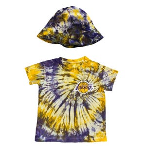 May include: A tie-dye baby outfit featuring a short-sleeved t-shirt and a bucket hat. The shirt and hat are in yellow, purple, and black tie-dye with the Los Angeles Lakers logo on the shirt.