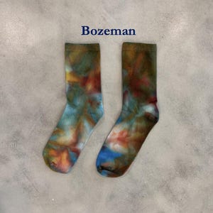 Puede incluir: Un par de calcetines con un patr&oacute;n de te&ntilde;ido anudado en tonos mezclados de azul, naranja, verde y marr&oacute;n. La palabra Bozeman est&aacute; impresa en azul oscuro sobre los calcetines. Los calcetines tienen un pu&ntilde;o acanalado y forma cl&aacute;sica.