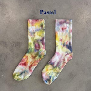 Puede incluir: Un par de calcetines de media ca&ntilde;a con un patr&oacute;n de te&ntilde;ido anudado en varios colores pastel, incluyendo amarillo, rosa, azul y verde. La palabra Pastel est&aacute; impresa en azul oscuro sobre los calcetines, que tienen un pu&ntilde;o acanalado y textura.