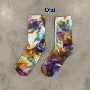 Puede incluir: Un par de calcetines de media ca&ntilde;a con un patr&oacute;n tie-dye multicolor con remolinos de color morado, amarillo, azul y blanco. La palabra Ojai est&aacute; impresa en azul oscuro sobre los calcetines, que tienen un pu&ntilde;o acanalado y aspecto suave.