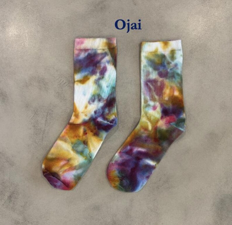 Puede incluir: Un par de calcetines de media ca&ntilde;a con un patr&oacute;n tie-dye multicolor con remolinos de color morado, amarillo, azul y blanco. La palabra Ojai est&aacute; impresa en azul oscuro sobre los calcetines, que tienen un pu&ntilde;o acanalado y aspecto suave.