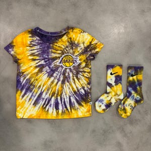 May include: A short-sleeve t-shirt and a pair of crew socks featuring a purple and yellow tie-dye pattern. The t-shirt displays the Los Angeles Lakers basketball team logo on the chest, while each sock features a small black Nike swoosh logo.