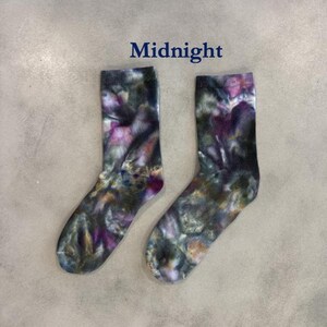 Puede incluir: Un par de calcetines de media ca&ntilde;a con un patr&oacute;n de te&ntilde;ido anudado en tonos verde oscuro, negro y morado. La palabra Midnight est&aacute; impresa en texto azul oscuro sobre los calcetines. Tienen un pu&ntilde;o acanalado y una estructura de tal&oacute;n est&aacute;ndar.