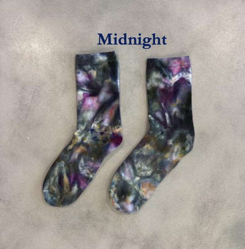 Puede incluir: Un par de calcetines de media ca&ntilde;a con un patr&oacute;n de te&ntilde;ido anudado en tonos verde oscuro, negro y morado. La palabra Midnight est&aacute; impresa en texto azul oscuro sobre los calcetines. Tienen un pu&ntilde;o acanalado y una estructura de tal&oacute;n est&aacute;ndar.