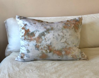 May include: A white pillow with a tie-dye pattern in shades of brown, orange, and blue. The pillow is lying on a white bed.