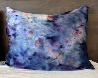 Puede incluir: Una almohada azul, morada y blanca con estampado tie-dye de tela suave y texturada. La almohada está sobre una cama blanca.