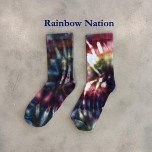 Puede incluir: Un par de calcetines de media ca&ntilde;a con un patr&oacute;n de te&ntilde;ido anudado en tonos rojo oscuro, azul oscuro, verde bosque y blanco. El texto Rainbow Nation aparece impreso en letras azul oscuro sobre los calcetines, que tienen un pu&ntilde;o acanalado.