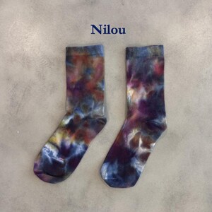 Puede incluir: Un par de calcetines de media ca&ntilde;a con un patr&oacute;n tie-dye en tonos mezclados de morado, azul, marr&oacute;n y amarillo. La palabra Nilou est&aacute; impresa en azul oscuro sobre los calcetines, que tienen un pu&ntilde;o acanalado y un aspecto suave y texturizado.