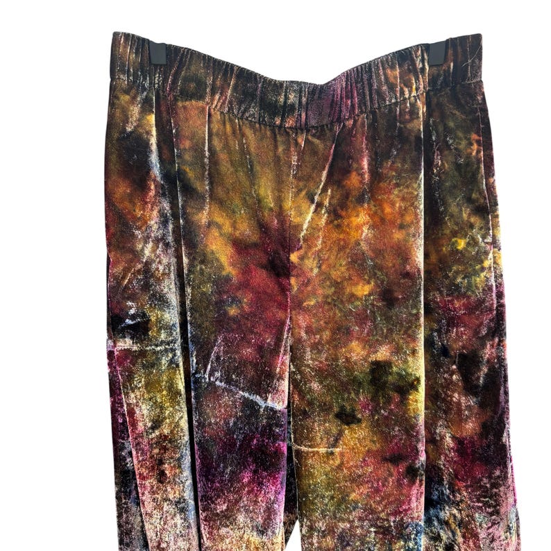 K&ouml;nnte beinhalten: Mehrfarbige Samthose mit elastischem Bund. Die Hose hat ein lebendiges Batikmuster in Braun-, Gelb-, Rot- und Lilat&ouml;nen. Der Stoff hat eine weiche, flie&szlig;ende Textur. Die Hose h&auml;ngt an einem schwarzen Kleiderb&uuml;gel.