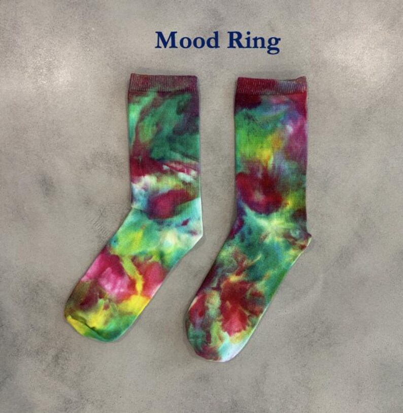 Puede incluir: Un par de calcetines de media ca&ntilde;a con un vibrante patr&oacute;n de te&ntilde;ido anudado en tonos mezclados de magenta, verde lima, amarillo y azul verdoso. El texto "Mood Ring" est&aacute; impreso en azul oscuro sobre los calcetines, que tienen un pu&ntilde;o acanalado.