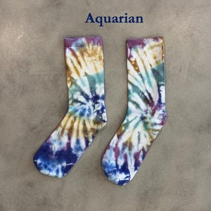 Puede incluir: Un par de calcetines con un patr&oacute;n de te&ntilde;ido anudado en tonos azul, morado, marr&oacute;n y blanco. La palabra Aquarian est&aacute; impresa en texto azul oscuro sobre los calcetines. Los calcetines tienen un pu&ntilde;o acanalado y un dise&ntilde;o en espiral.
