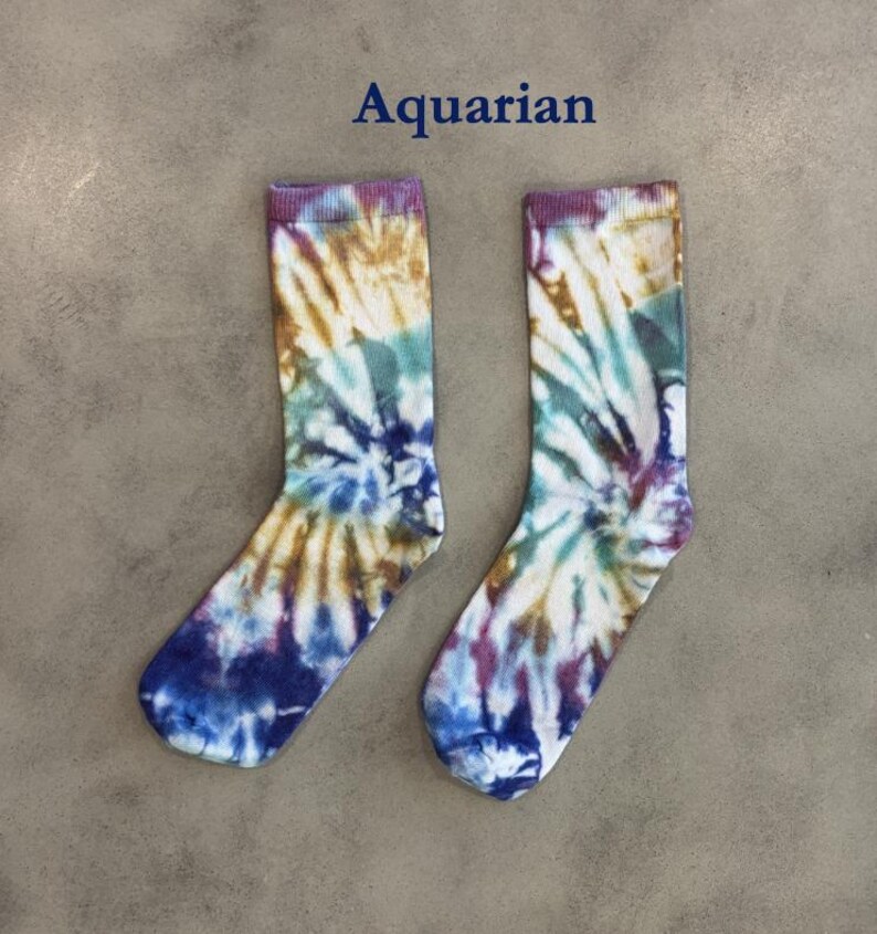 Puede incluir: Un par de calcetines con un patr&oacute;n de te&ntilde;ido anudado en tonos azul, morado, marr&oacute;n y blanco. La palabra Aquarian est&aacute; impresa en texto azul oscuro sobre los calcetines. Los calcetines tienen un pu&ntilde;o acanalado y un dise&ntilde;o en espiral.