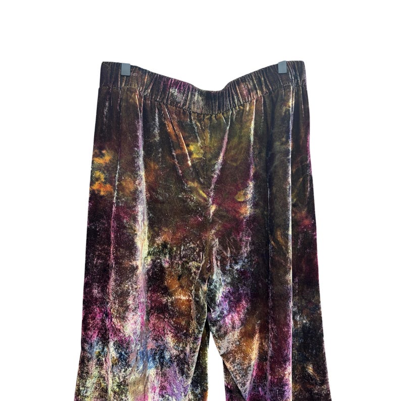 K&ouml;nnte beinhalten: Samthose mit elastischem Bund, mit einem lebendigen Batikmuster in Braun-, Lila-, Gelb- und Blaut&ouml;nen. Die Hose h&auml;ngt vor einem wei&szlig;en Hintergrund. Der Stoff hat ein weiches, flie&szlig;endes Aussehen.