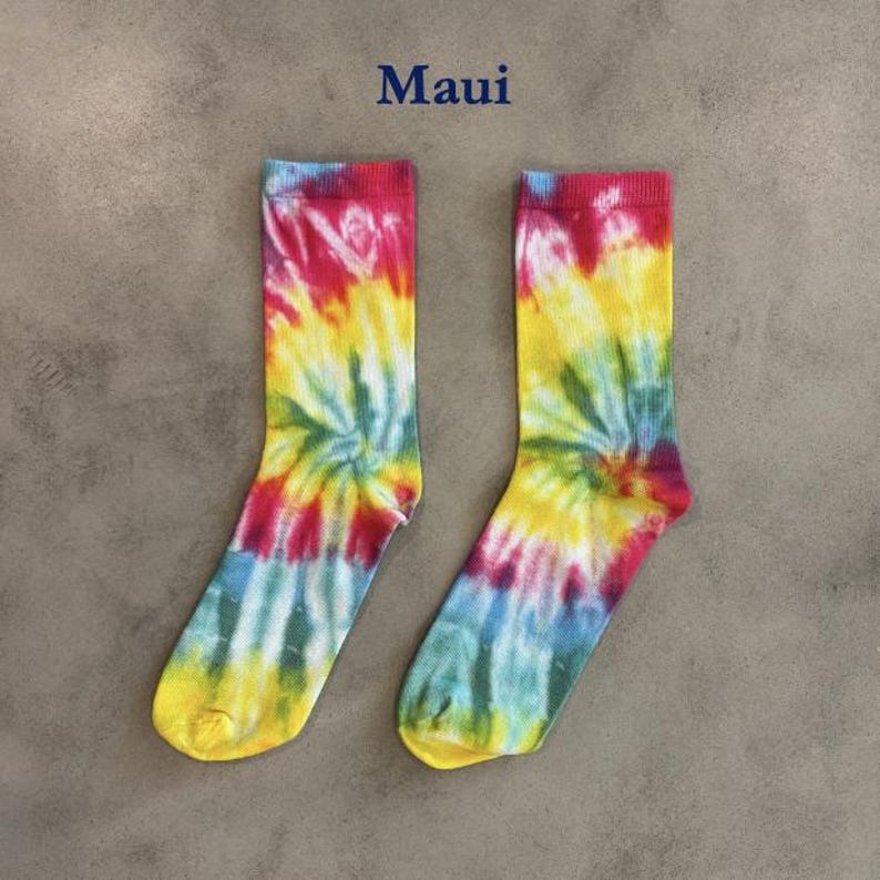 Puede incluir: Un par de calcetines con un vibrante patr&oacute;n tie-dye en tonos amarillo, rosa, azul y verde. La palabra Maui est&aacute; impresa en azul oscuro sobre los calcetines. Los calcetines tienen un pu&ntilde;o acanalado y una textura de punto est&aacute;ndar.