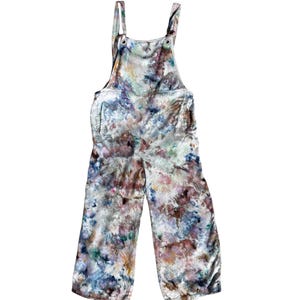 Può includere: Salopette tie-dye con un motivo simile a un acquerello. L'indumento presenta spalline regolabili, una tasca a pettorina e tasche laterali. I colori includono sfumature di blu, viola, marrone e bianco, creando un look unico e artistico. La salopette è realizzata in tessuto morbido.