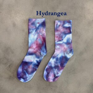 Puede incluir: Un par de calcetines de media ca&ntilde;a con un patr&oacute;n de te&ntilde;ido anudado en tonos azul, morado y blanco. La palabra Hydrangea est&aacute; impresa en texto azul oscuro sobre los calcetines. Los calcetines tienen un pu&ntilde;o acanalado y un aspecto suave.