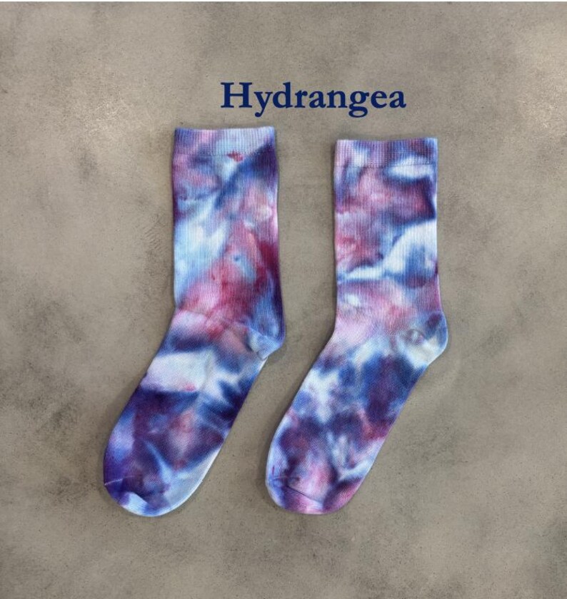 Puede incluir: Un par de calcetines de media ca&ntilde;a con un patr&oacute;n de te&ntilde;ido anudado en tonos azul, morado y blanco. La palabra Hydrangea est&aacute; impresa en texto azul oscuro sobre los calcetines. Los calcetines tienen un pu&ntilde;o acanalado y un aspecto suave.